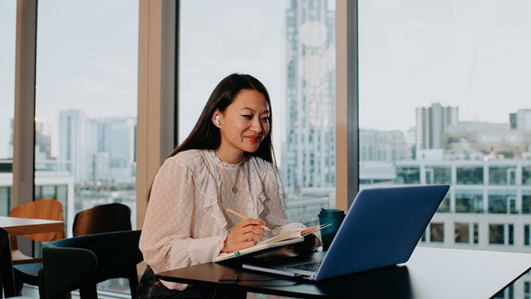 A content and confident young woman sits behind a laptop, taking notes.