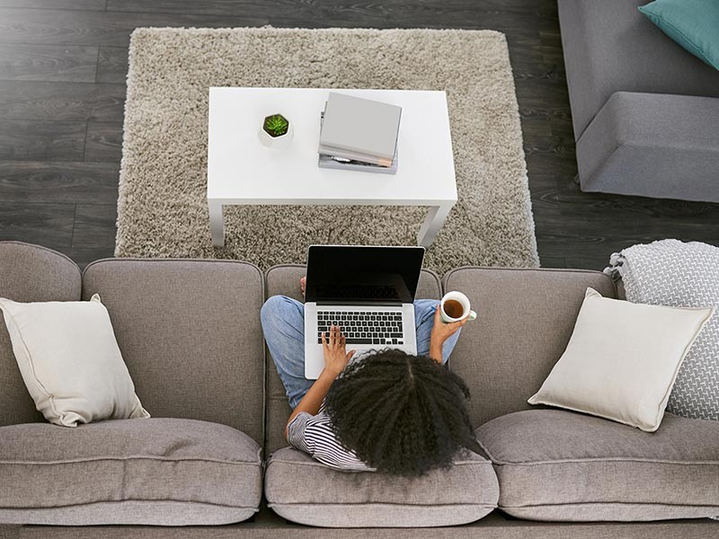 High angle shot of a young woman using a laptop and having coffee on the sofa at home